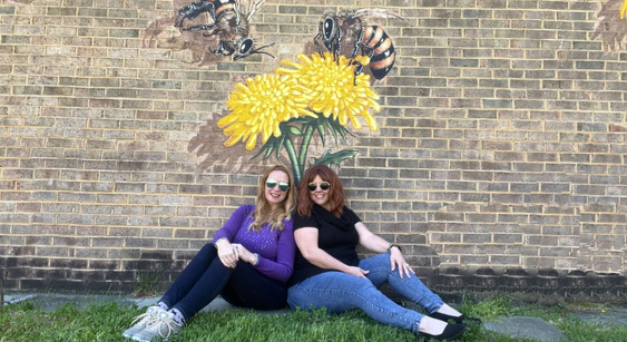 Denise and Cheryl sitting on the grass in front of a mural with bees on it